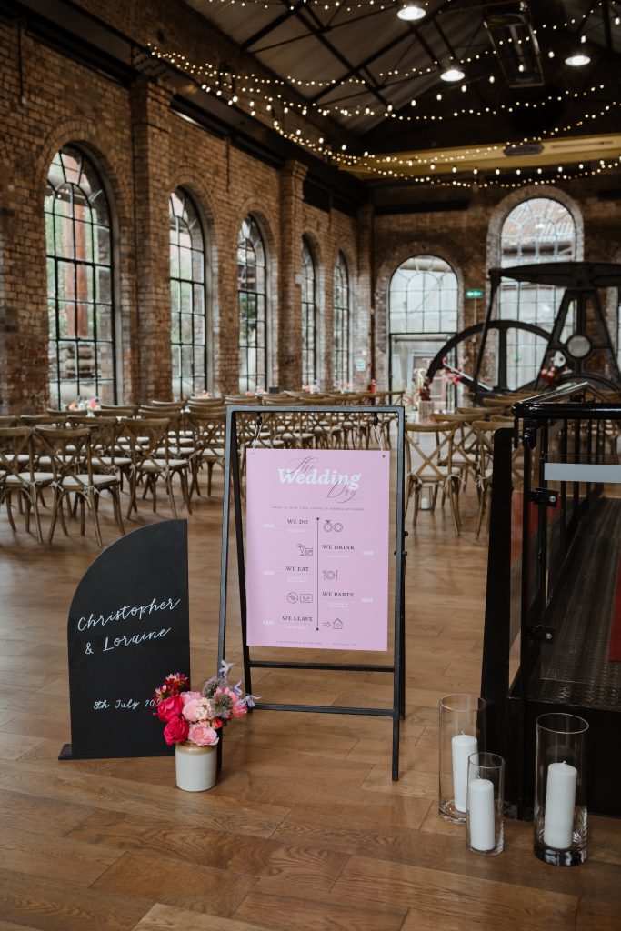 A Scottish wedding venue in the national mining museum with wedding decoration containing chairs in the background and a signage in front with the wedding timeline and the name of the couple.
